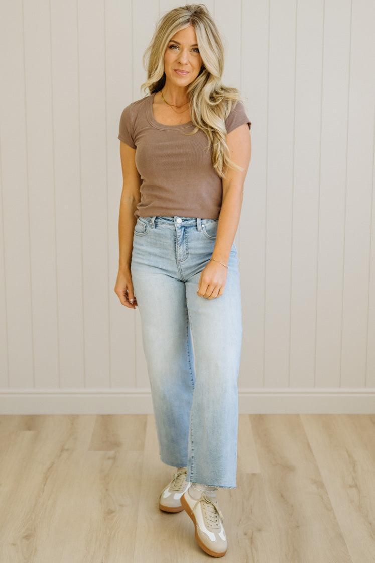 Woman wearing a brown top and light blue jeans standing against a white paneled wall.