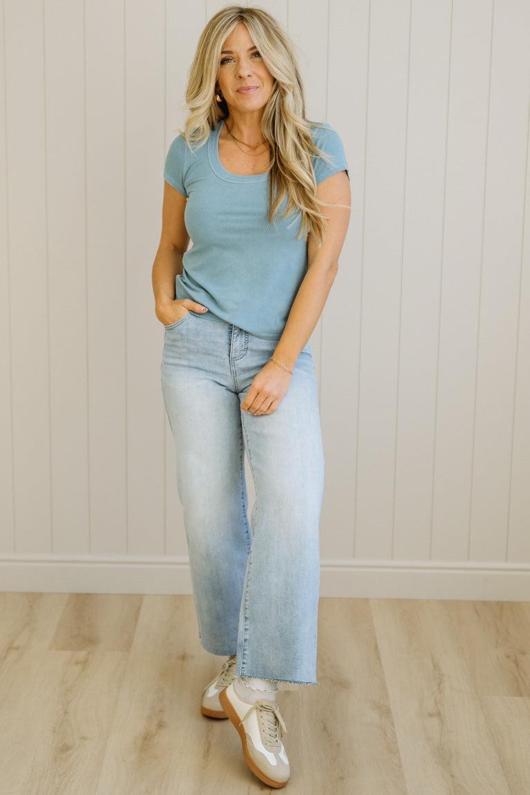 Woman wearing a light blue top and jeans standing against a white paneled wall.