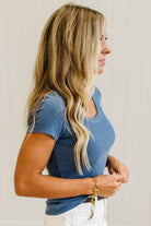A woman with long blonde hair is shown in profile wearing a short-sleeve blue scoop-neck t-shirt and white pants, accessorized with a gold bracelet against a light neutral background.