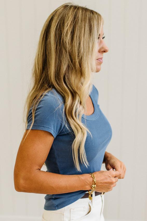A woman with long blonde hair is shown in profile wearing a short-sleeve blue scoop-neck t-shirt and white pants, accessorized with a gold bracelet against a light neutral background.