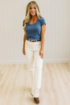 A woman modeling a blue short-sleeve scoop-neck t-shirt paired with off-white high-waisted straight-leg jeans, a thin black belt, and brown boots, standing in a room with light wood floors and white paneled walls.