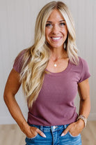 A smiling blonde woman wearing a mauve-purple short-sleeve crewneck t-shirt and blue jeans, standing against a light-colored paneled wall.