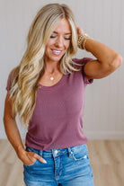 A woman with long blonde hair wearing a mauve-purple short-sleeve scoop neck t-shirt and blue jeans, posing in a brightly lit indoor setting.