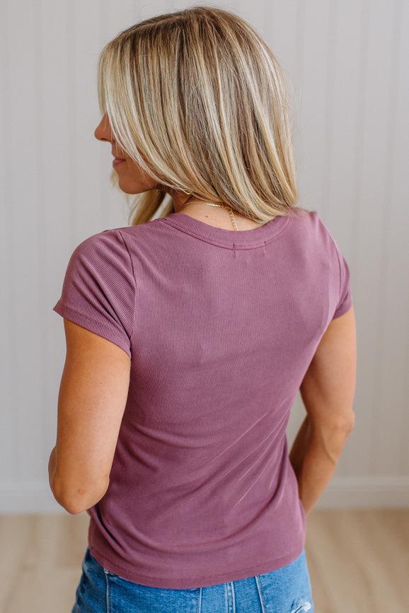 A woman with blonde hair is shown from the back wearing a short-sleeve crewneck t-shirt in a dusty mauve or plum color, paired with blue jeans, set against a light-colored paneled wall and wood floor.