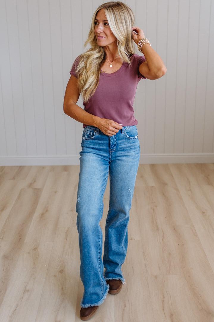 A woman modeling a mauve short-sleeve t-shirt and light-wash blue flare jeans with brown boots, standing on a light wood floor against a white paneled wall.