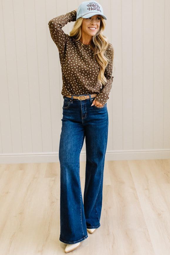 Blonde woman in dark jeans and a light denim ball cap wearing a brown long sleeve tee with botanical print.