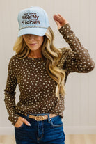 Blonde woman in dark jeans and a light denim ball cap wearing a brown long sleeve tee with botanical print.