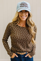 Blonde woman in dark jeans and a light denim ball cap wearing a brown long sleeve tee with botanical print.