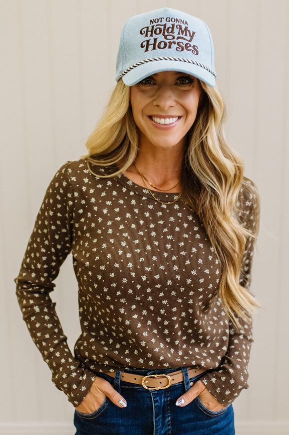 Blonde woman in dark jeans and a light denim ball cap wearing a brown long sleeve tee with botanical print.
