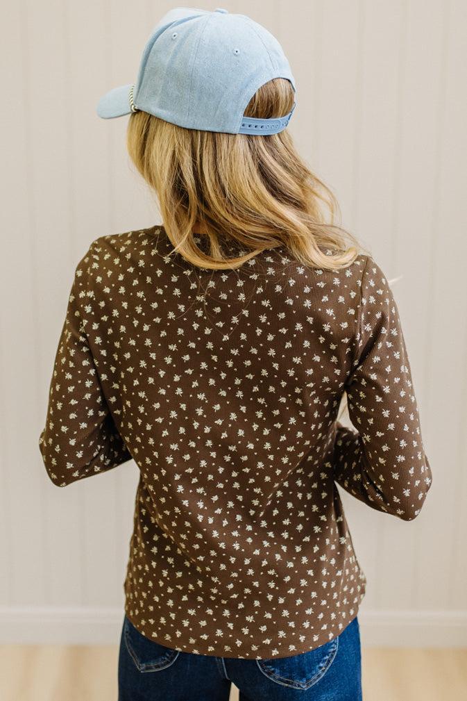 Blonde woman in dark jeans and a light denim ball cap wearing a brown long sleeve tee with botanical print.
