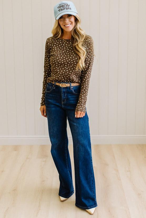 Blonde woman in dark jeans and a light denim ball cap wearing a brown long sleeve tee with botanical print.