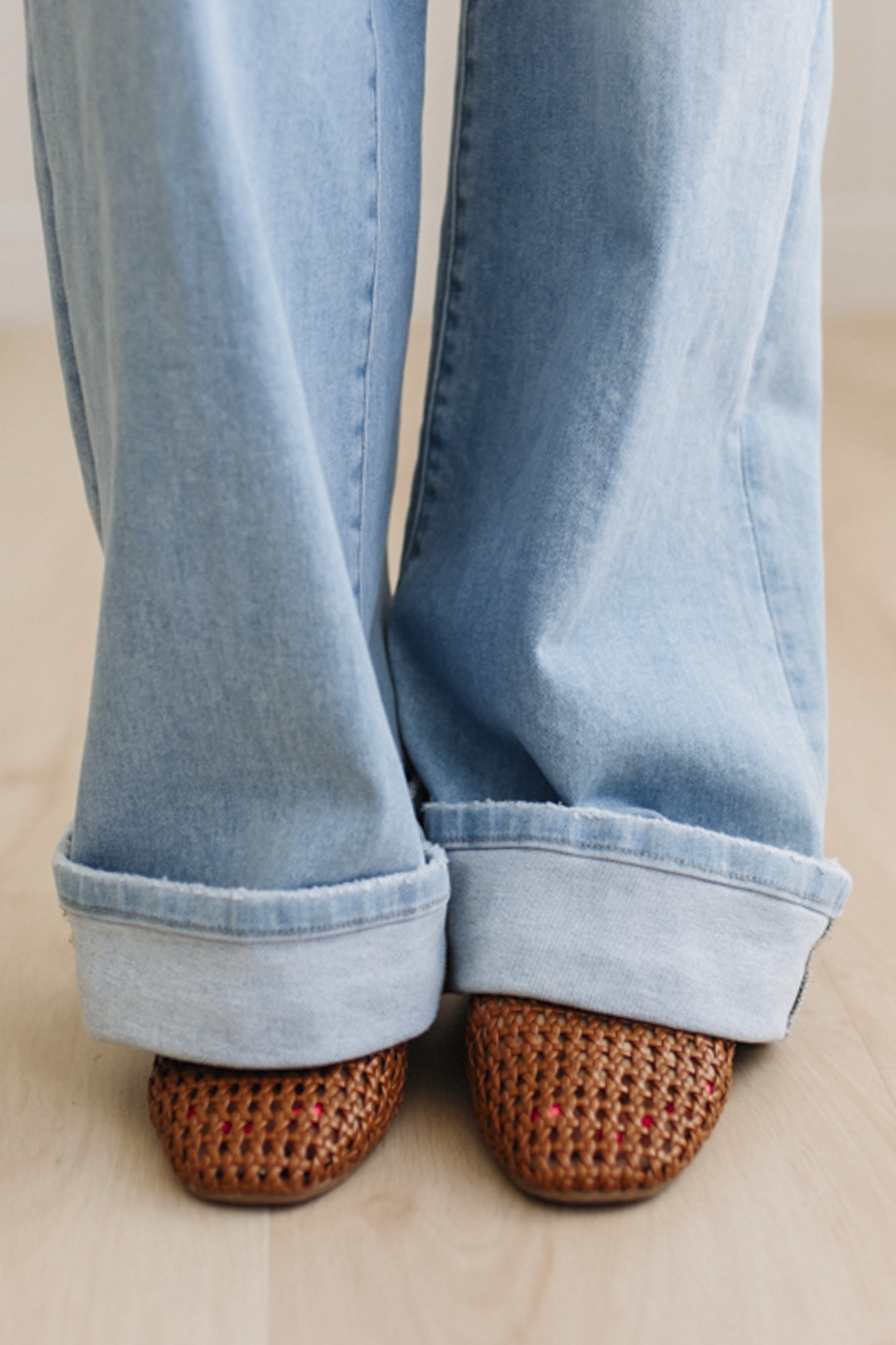 Light blue jeans with rolled-up cuffs worn with brown woven slippers on a beige surface.