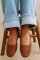 Brown woven flats worn with rolled-up jeans on a wooden chair.