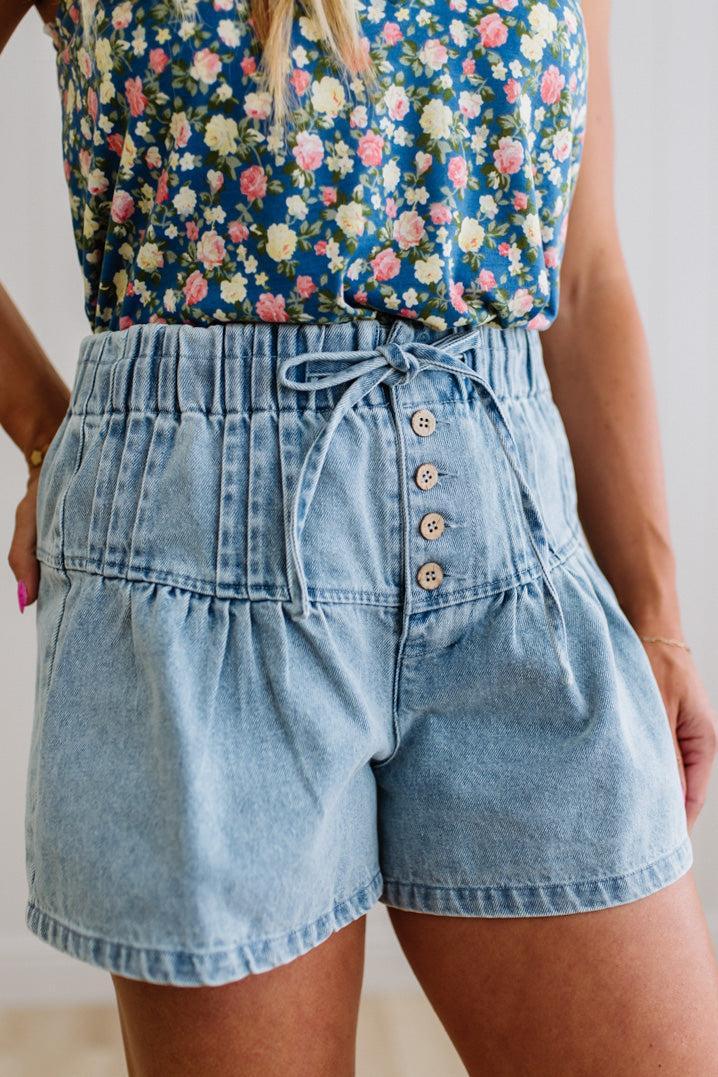 Blonde woman in a blue floral tank wearing drawstring waist flare denim shorts, front button up, with pintuck detail, and light washed denim fabric.