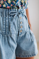 Blonde woman in a blue floral tank wearing drawstring waist flare denim shorts, front button up, with pintuck detail, and light washed denim fabric.