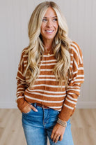 A smiling blonde woman wearing a brown and cream striped knit zip-up sweater and blue jeans, standing against a light-colored paneled wall and wood floor.