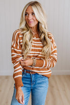 A woman with long blonde hair wearing a rust-orange and white striped knit sweater, blue jeans, and gold bracelets, standing in a brightly lit room with light wood floors and white paneled walls.