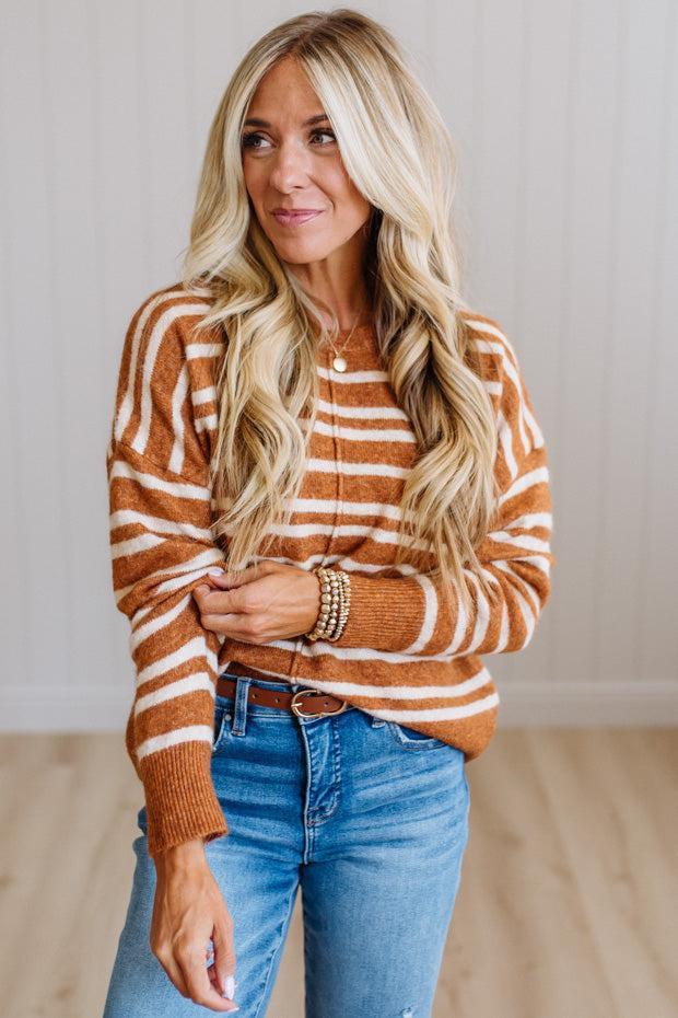 A woman with long blonde hair wearing a rust-orange and white striped knit sweater, blue jeans, and gold bracelets, standing in a brightly lit room with light wood floors and white paneled walls.