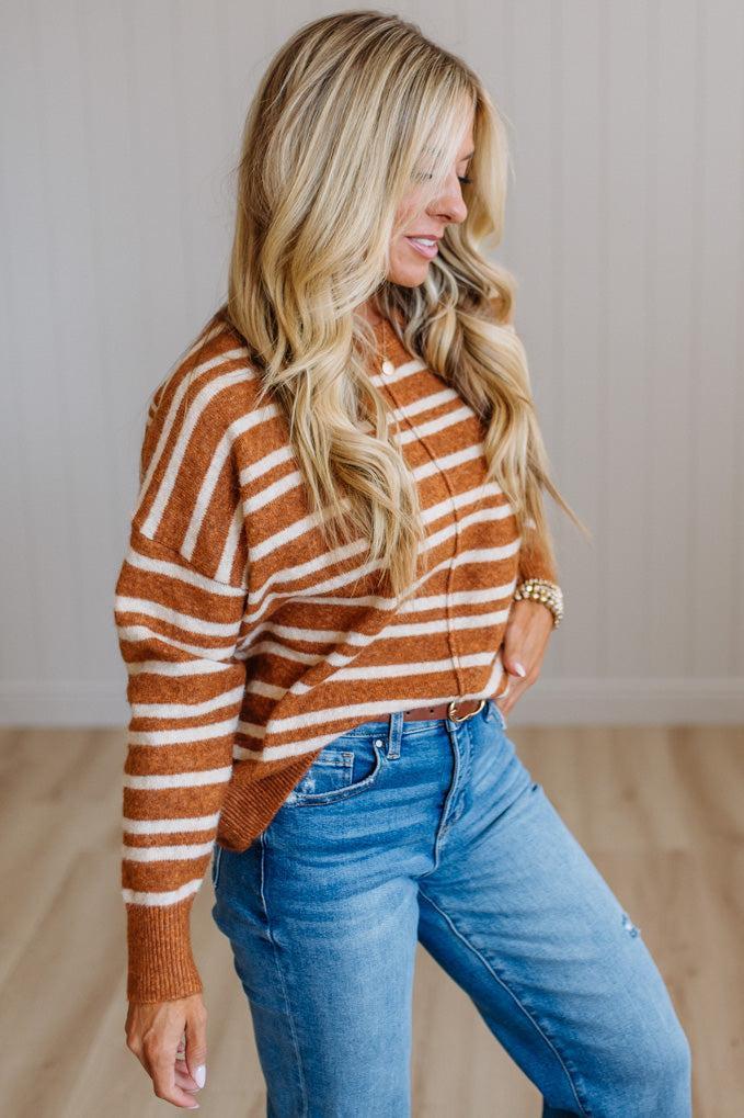 A woman wearing a rust-orange and white striped knit sweater with blue jeans, standing in a room with light wood floors and a neutral paneled wall.