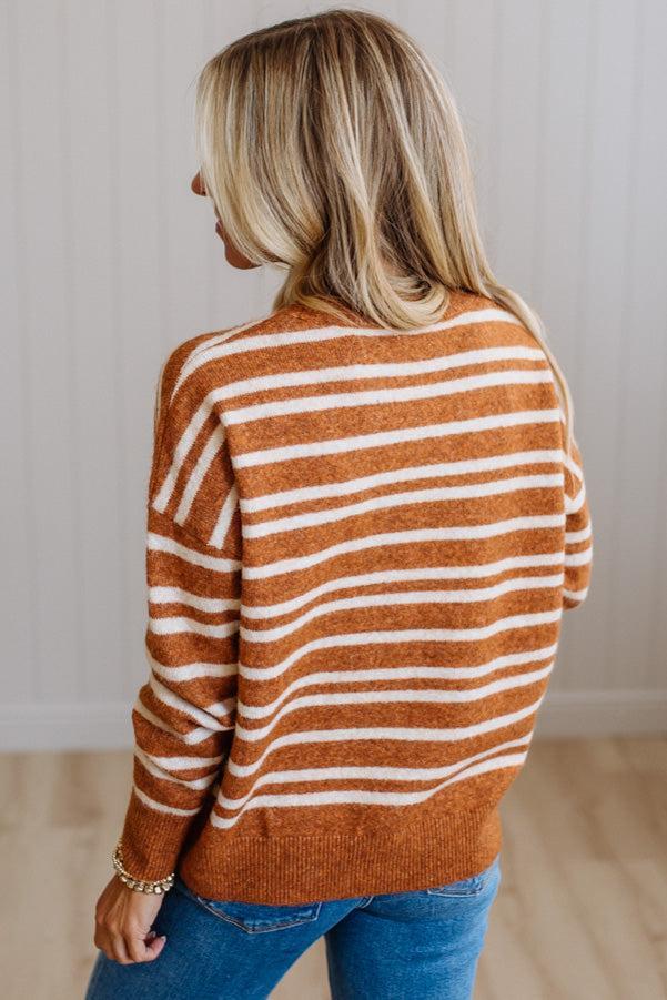 A woman shown from the back wearing a rust-orange and white horizontally striped knit sweater with blue jeans, standing in a brightly lit indoor setting.
