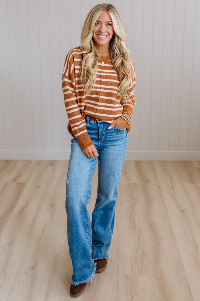 A woman stands in a room with light wood floors and white paneled walls, wearing a rust-orange and white striped long-sleeve knit top, blue jeans, and brown boots.