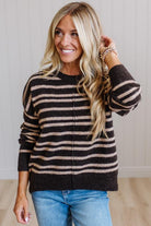 A smiling blonde woman wearing a dark brown and beige horizontally striped knit sweater with a front zipper, paired with blue jeans, standing against a light wood-paneled wall.