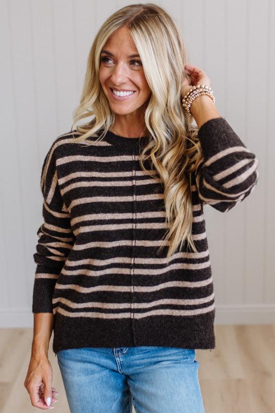 A smiling blonde woman wearing a dark brown and beige horizontally striped knit sweater with a front zipper, paired with blue jeans, standing against a light wood-paneled wall.