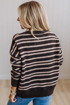 A woman with blonde hair is shown from behind wearing a dark brown and beige horizontally striped knit sweater and blue jeans, standing against a white paneled wall.