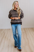 A woman modeling a dark brown and beige striped knit sweater with blue jeans and brown boots, standing in a room with light wood floors and white paneled walls.