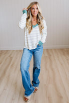 A woman modeling a white long-sleeve striped knit top with teal trim and blue jeans, standing on a light wood floor against a white paneled wall.