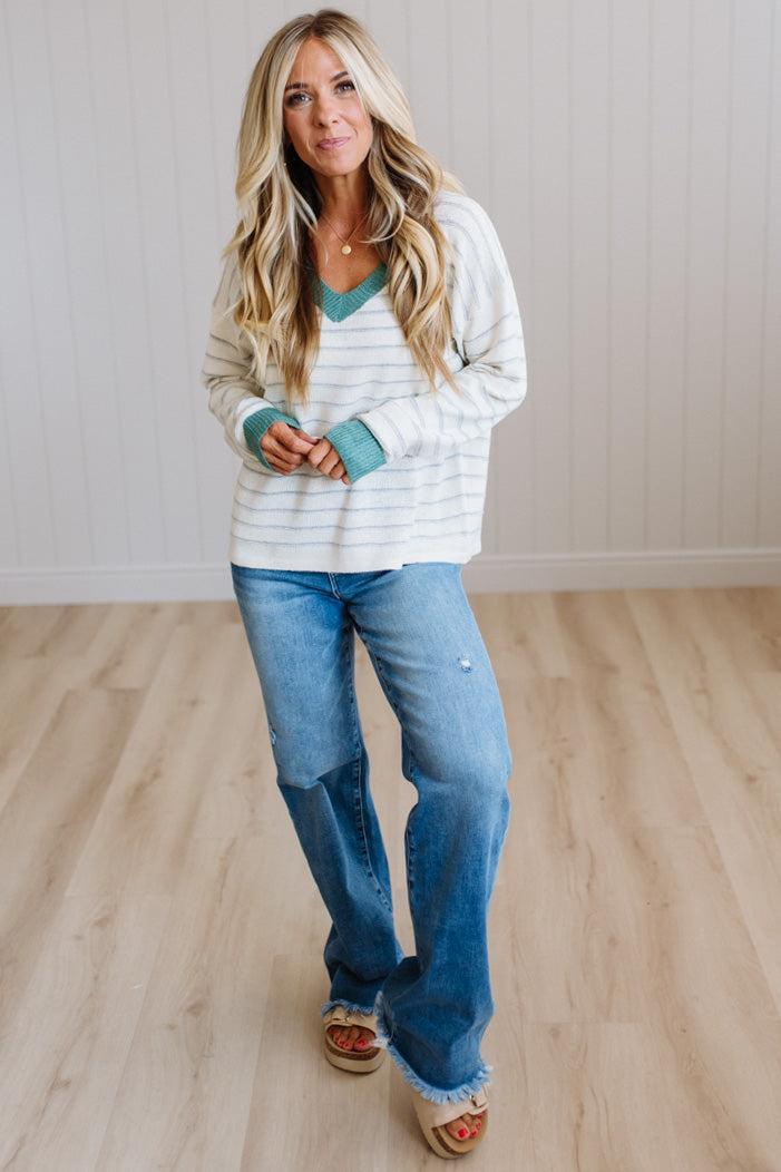 A woman modeling a white and light blue striped long-sleeve V-neck top with teal trim, paired with blue jeans and tan slide sandals, standing in a room with light wood floors and white paneled walls.