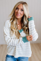A woman with long blonde hair wearing a white and light blue striped knit sweater with teal green trim on the collar and cuffs, paired with blue jeans, standing in a brightly lit indoor setting.