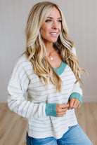 A woman with long blonde hair wearing a white and light blue striped knit sweater with teal green trim, paired with blue jeans, standing in a brightly lit room.