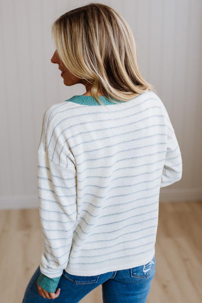 A woman shown from the back wearing a cream and gray striped knit sweater with a teal ribbed collar and cuffs, paired with blue jeans in a brightly lit indoor room.