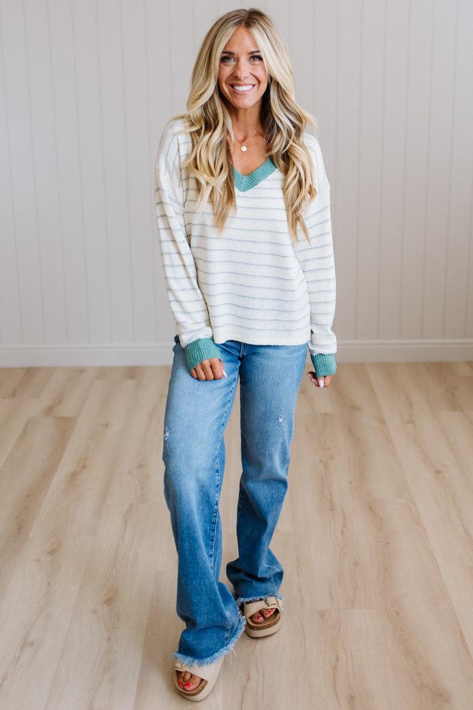 A woman stands in a bright room with light wood floors and white paneled walls, wearing a white long-sleeve V-neck top with thin teal stripes and teal cuffs, paired with blue jeans and tan sandals.