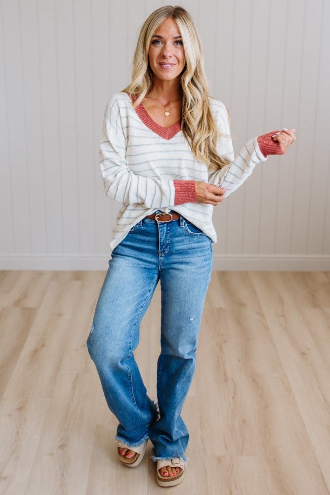 A woman modeling a white and gray striped long-sleeve V-neck sweater with coral pink trim, paired with blue jeans and brown sandals, standing in a room with light wood floors and white paneled walls.