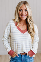 A smiling blonde woman wearing a cream and gray striped V-neck sweater with rust-colored trim, paired with blue jeans, standing against a light neutral wall.