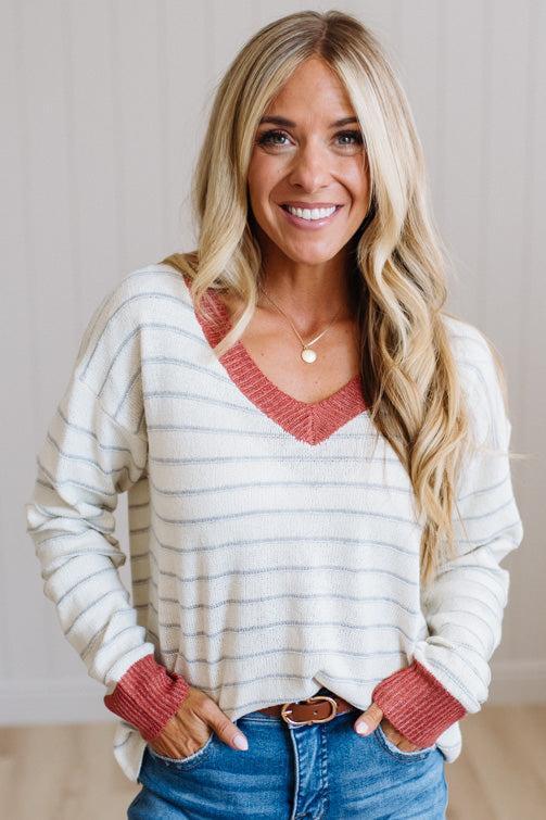 A smiling blonde woman wearing a cream and gray striped V-neck sweater with rust-colored trim, paired with blue jeans, standing against a light neutral wall.