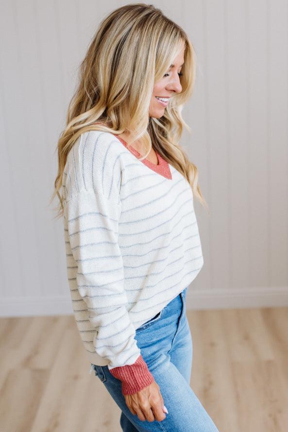 A woman wearing a white long-sleeve knit sweater with thin gray horizontal stripes, layered over a coral pink V-neck top, paired with blue jeans, standing in a brightly lit room with light wood floors and white paneled walls.