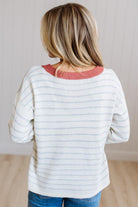 A woman viewed from behind wearing a cream-colored knit sweater with thin light blue horizontal stripes and a contrasting dusty rose/coral crew neck, paired with blue jeans.