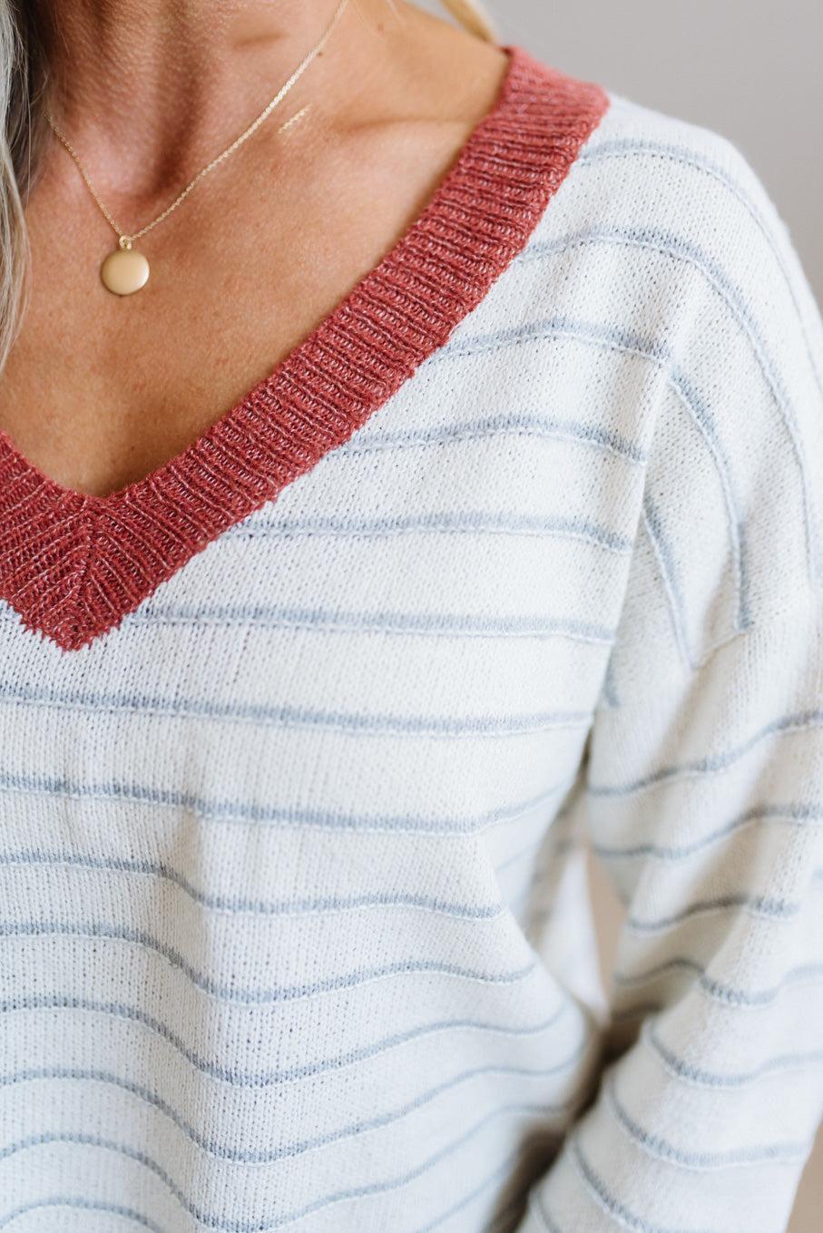 Close-up of a person wearing a ribbed V-neck knit sweater with thin horizontal light blue stripes and a rust-red ribbed collar, paired with a delicate gold necklace.