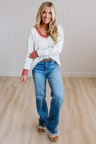 A woman modeling a white and gray striped long-sleeve henley shirt with red cuffs, paired with blue jeans and tan sandals, standing in a room with light wood floors and white paneled walls.
