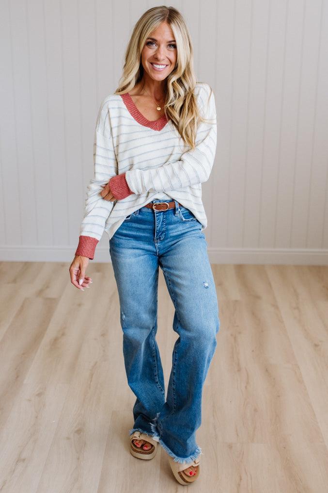 A woman modeling a white and gray striped long-sleeve henley shirt with red cuffs, paired with blue jeans and tan sandals, standing in a room with light wood floors and white paneled walls.