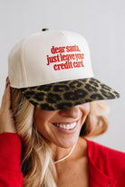 Blonde woman in a red top and gold necklace wearing a cream hat with leopard brim and red embroidered text that says "dear santa just leave your credit card"