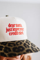 Blonde woman in a red top and gold necklace wearing a cream hat with leopard brim and red embroidered text that says "dear santa just leave your credit card"