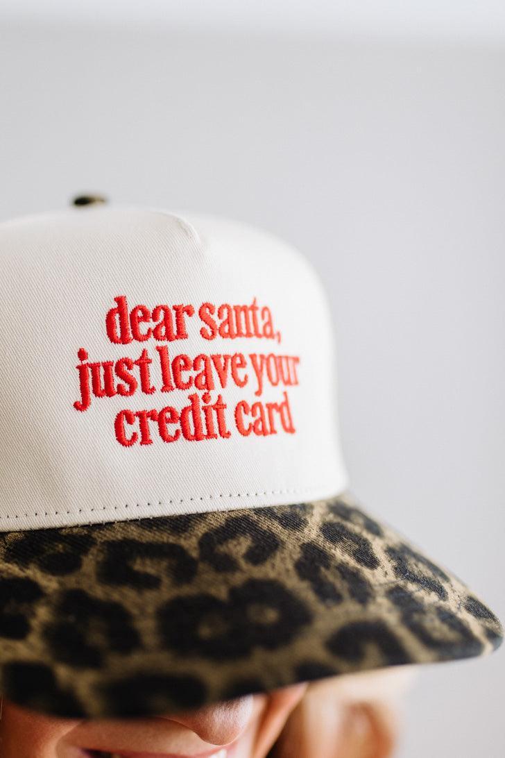 Blonde woman in a red top and gold necklace wearing a cream hat with leopard brim and red embroidered text that says "dear santa just leave your credit card"