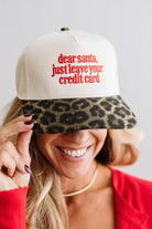 Blonde woman in a red top and gold necklace wearing a cream hat with leopard brim and red embroidered text that says "dear santa just leave your credit card"