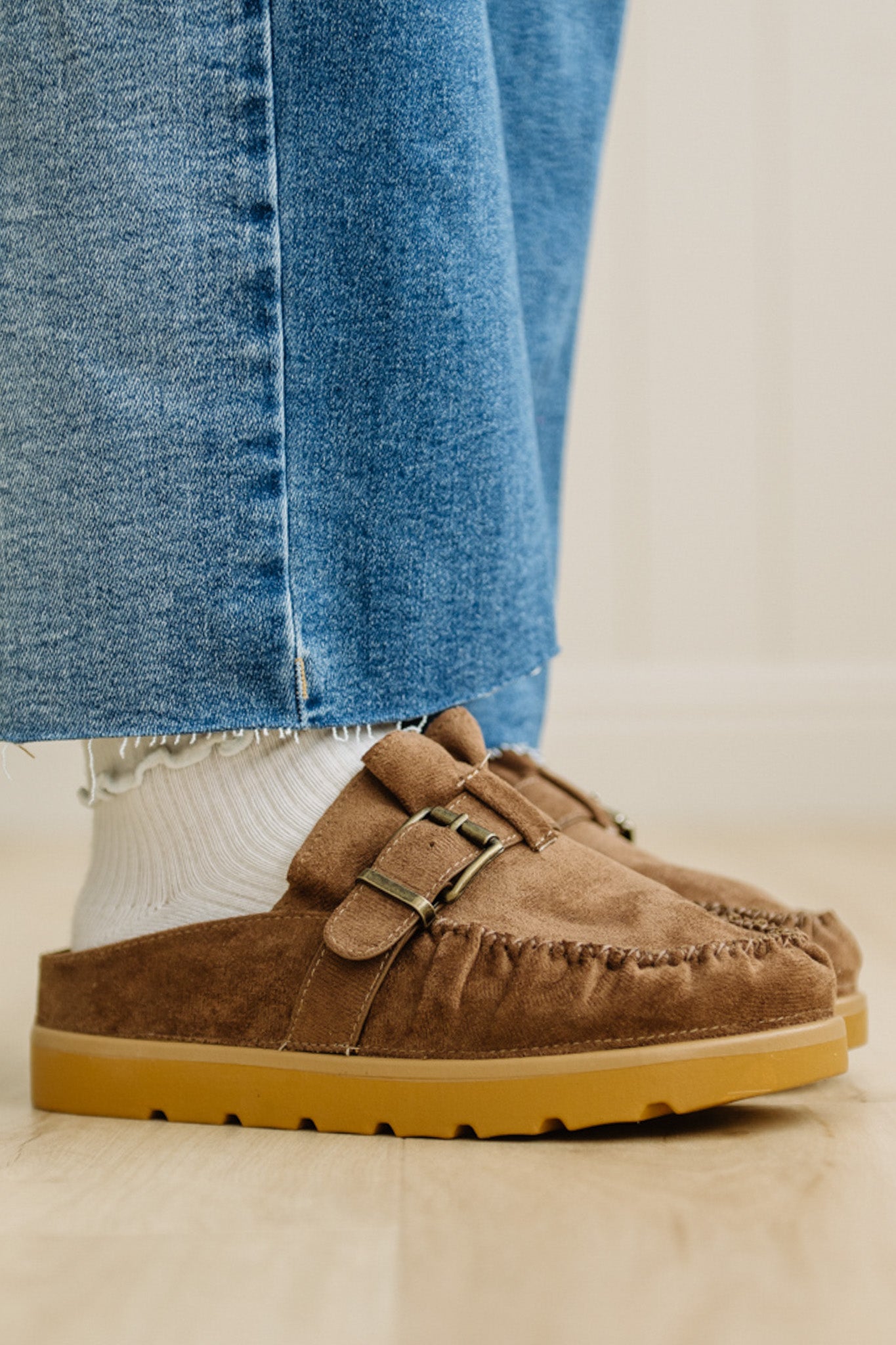 Brown suede shoes with a buckle worn with blue jeans and white socks on a light background