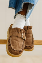 Brown suede loafers worn with ripped jeans and white socks on a neutral background
