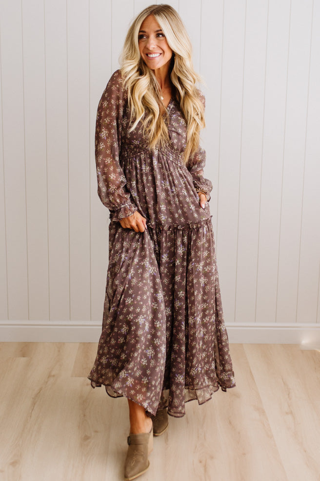 A woman modeling a long-sleeve, tiered maxi dress with a brown and beige floral pattern, standing in front of a white paneled wall and light wood floor.